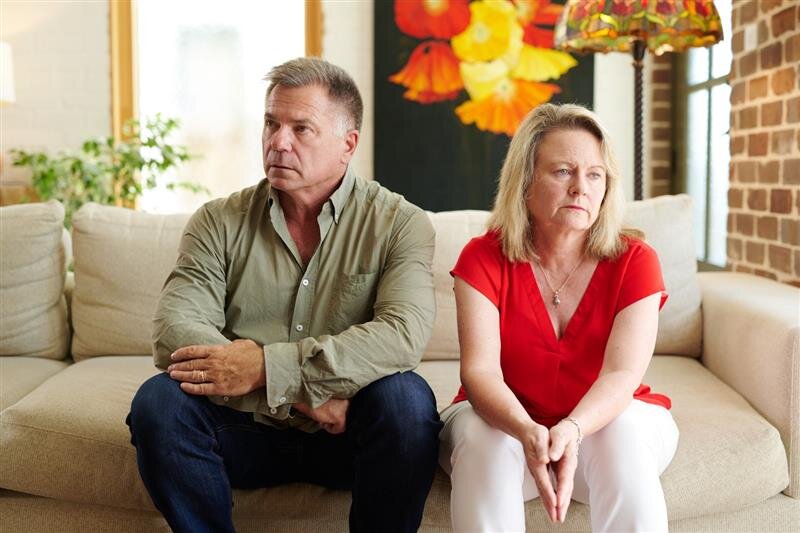 A man and woman sit on a couch in a living room, both looking away from each other with serious expressions